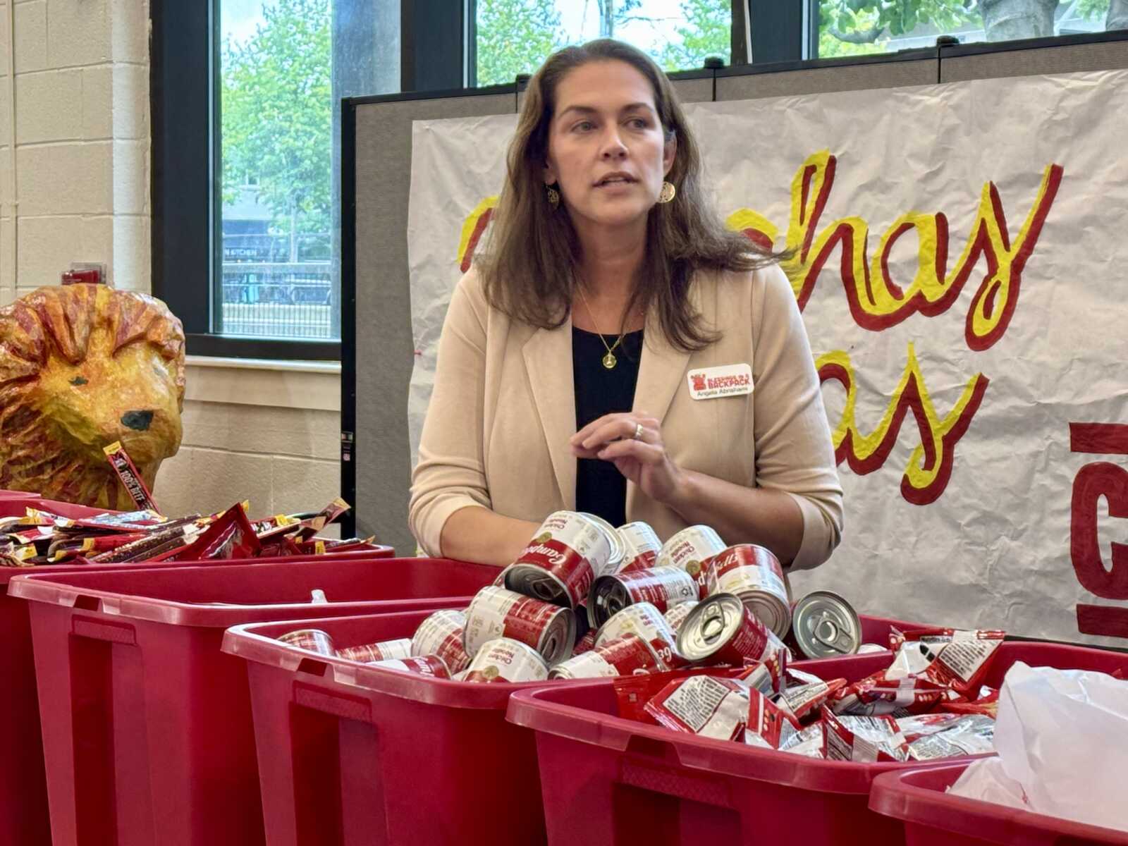 Volunteer in a beige blazer stands behind red bins filled with canned food at a donation drive.