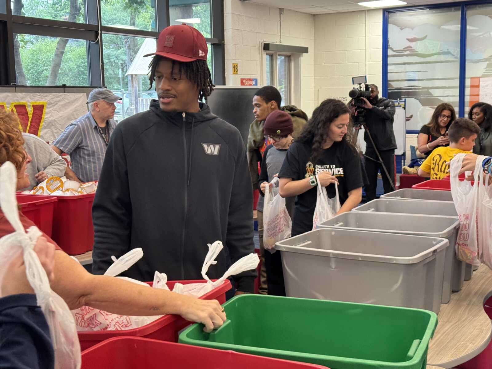 Volunteer in a black hoodie and red cap sorts bags at a community event with volunteers and a photographer in the background.