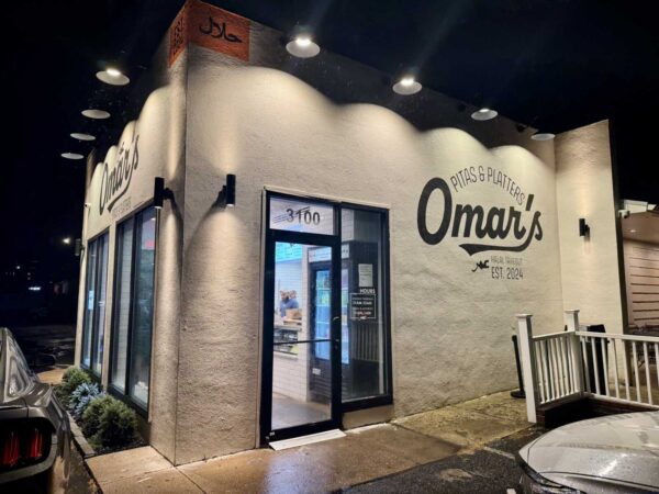 Night exterior of Omar's Pitas & Platters halal takeout shop, bold logo on beige stucco wall under bright warm lighting