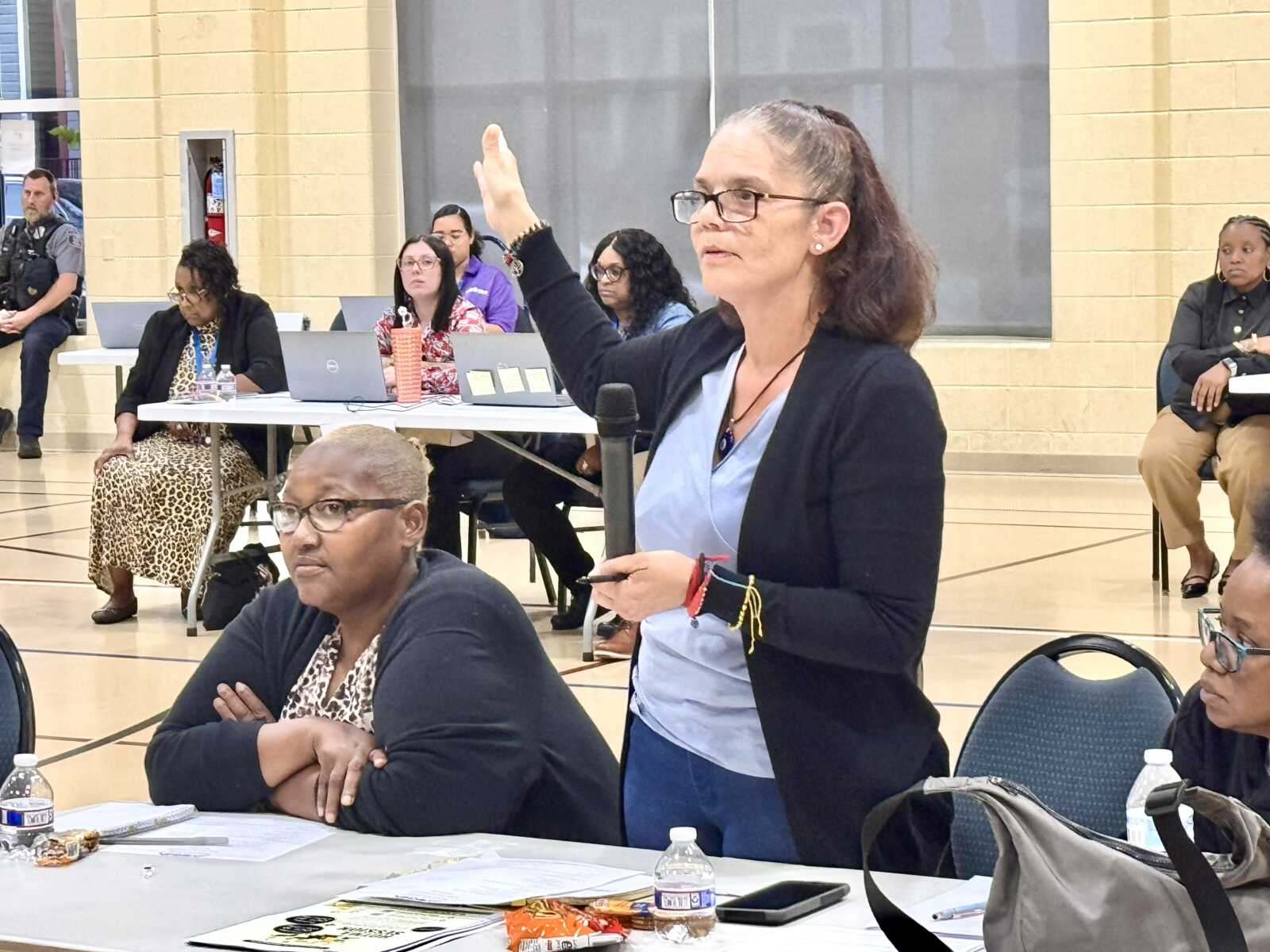 Woman standing with microphone, raising her hand to speak at a community meeting in a gymnasium. Lead panel and attendees sit at tables with laptops in the background.