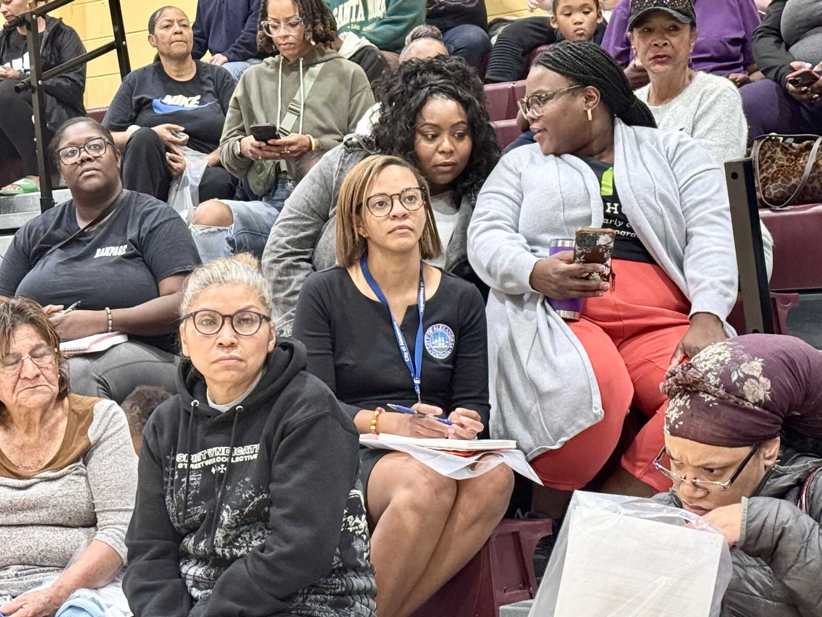Group of diverse people seated in gym bleachers listening at an event; woman with lanyard takes notes in front center.