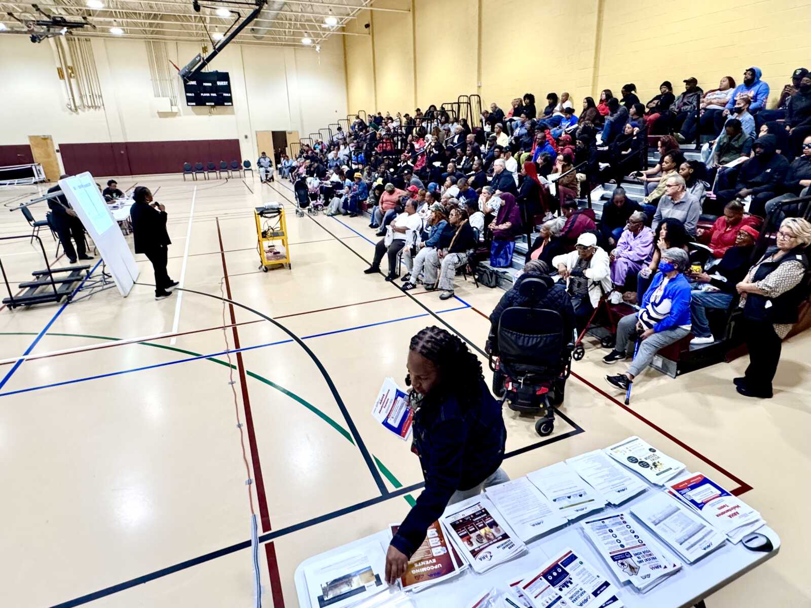 Speaker presenting to a diverse audience in a gymnasium, with a whiteboard, projector, and a table of brochures in the foreground.