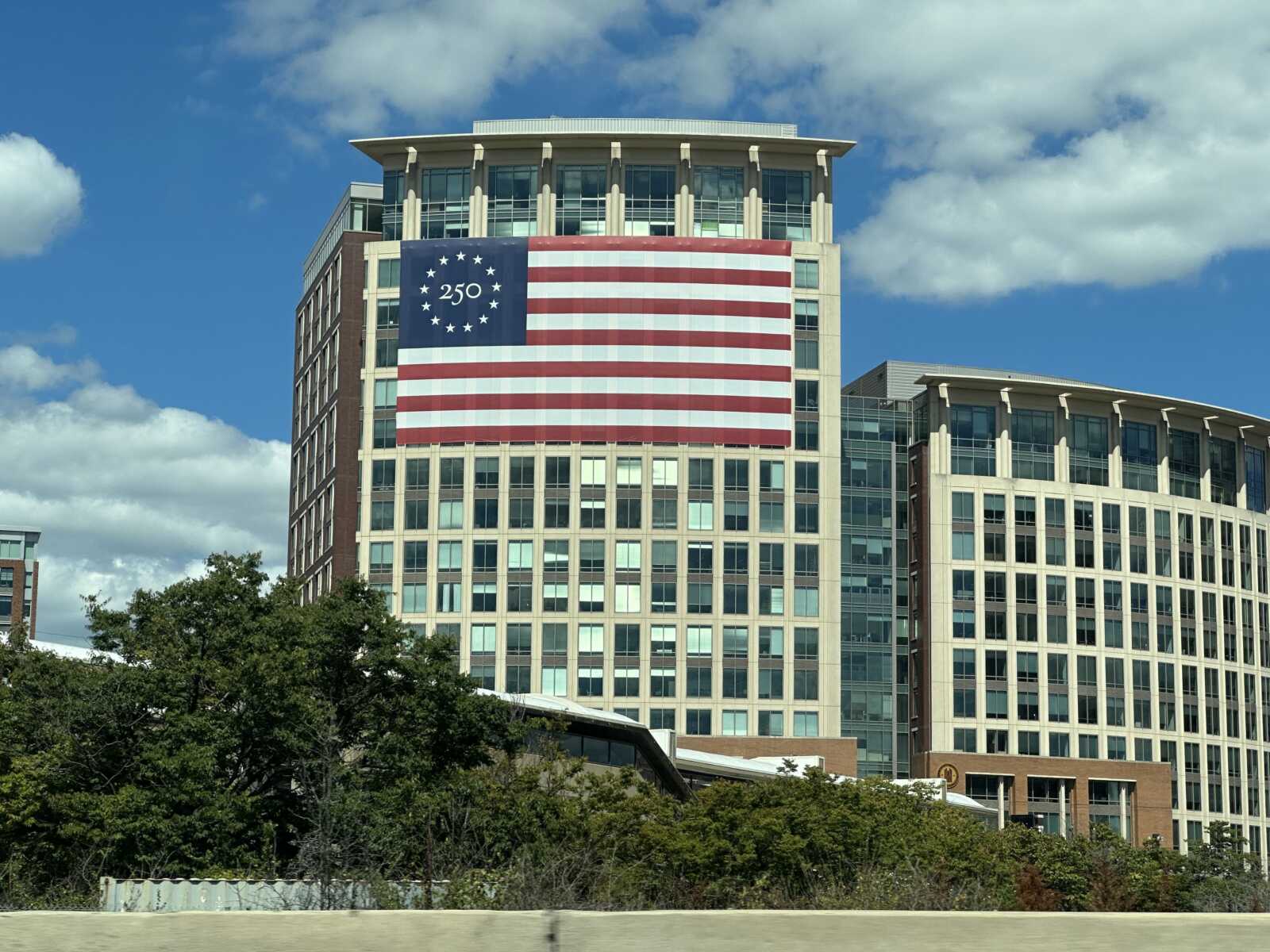 IMG_6505 morning notes | ALXnow Modern office building with a large American flag banner covering its center, featuring a blue field with'250' and stars, under a blue sky with clouds