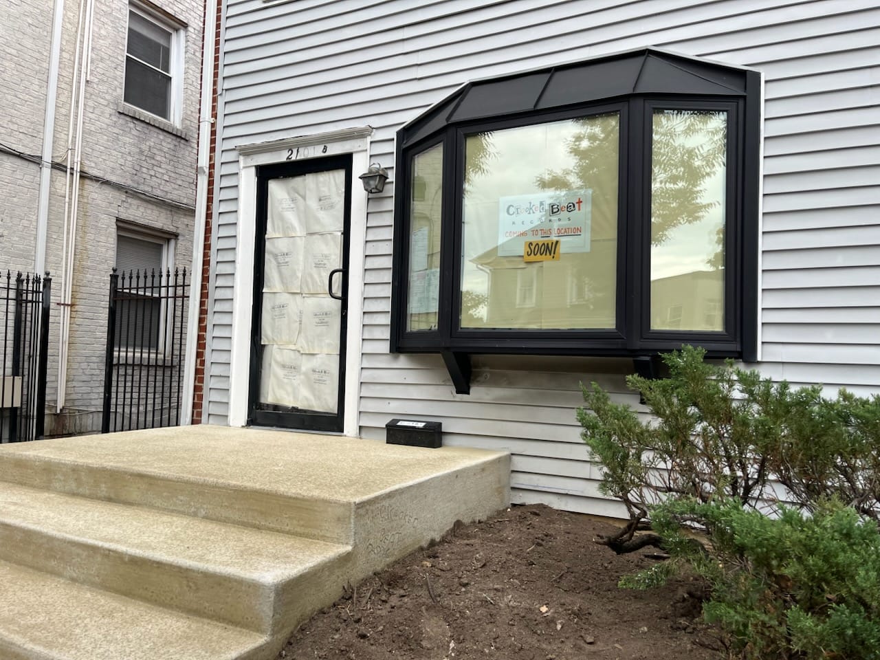 Building entrance with a black-framed door boarded over and a large bay window to the right; sign reads 'Coming to this location soon!' (under construction)