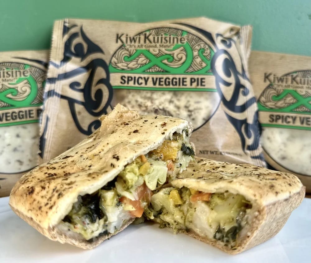 Cross-section of a baked spicy veggie pie showing diced peppers, corn, spinach and cheese filling, with Kiwi Cuisine Spicy Veggie Pie packages in the background.