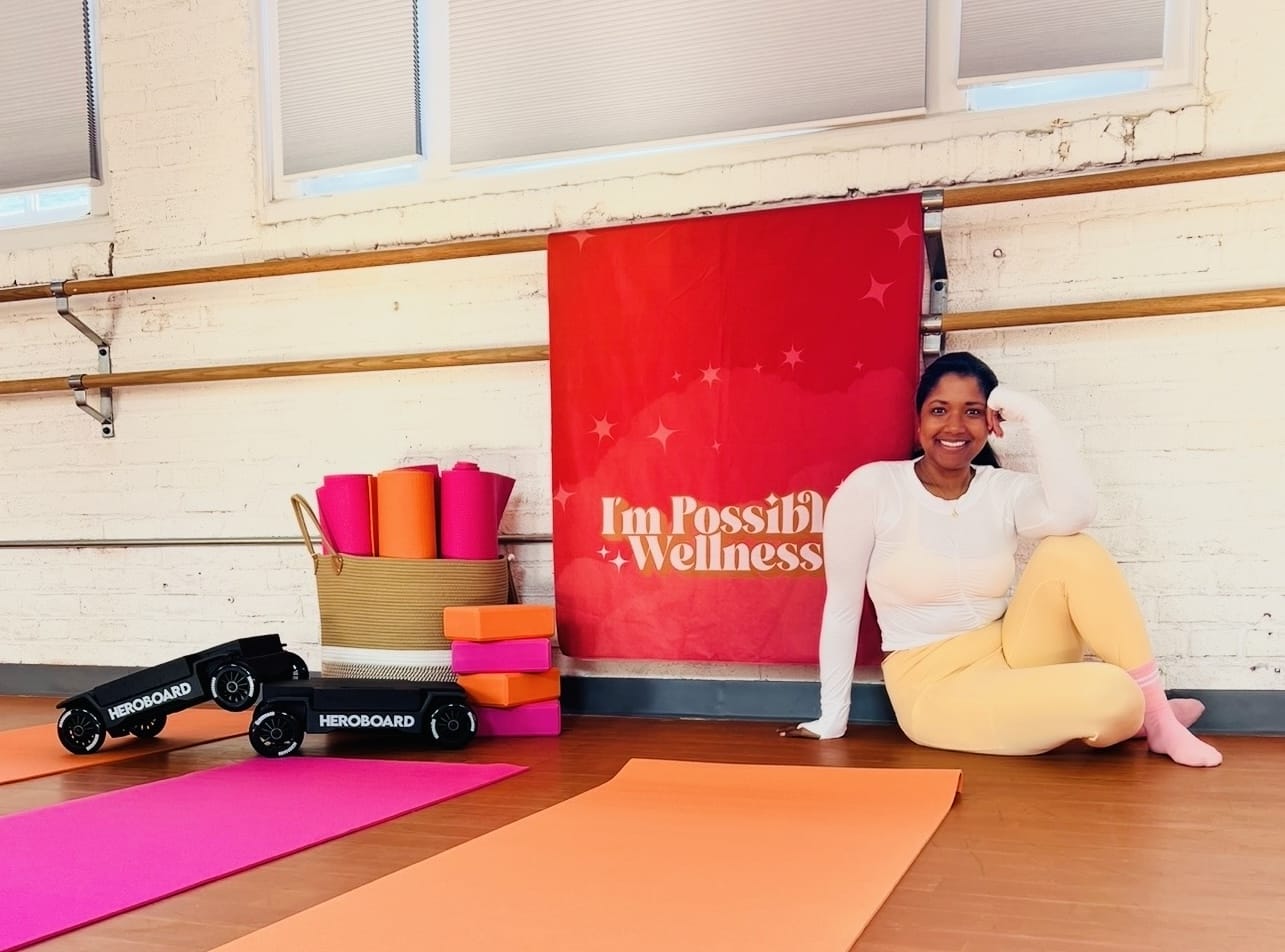 Una mujer sonriente se sienta en el suelo de un estudio de bienestar junto a colchonetas de yoga de color naranja y rosa, bloques y una pancarta roja que dice 