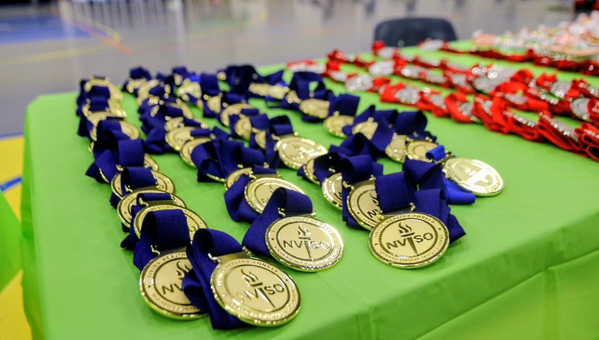 Rows of gold medals with blue ribbons lined up on a bright green table in a gym, ready for award distribution (NVSO).