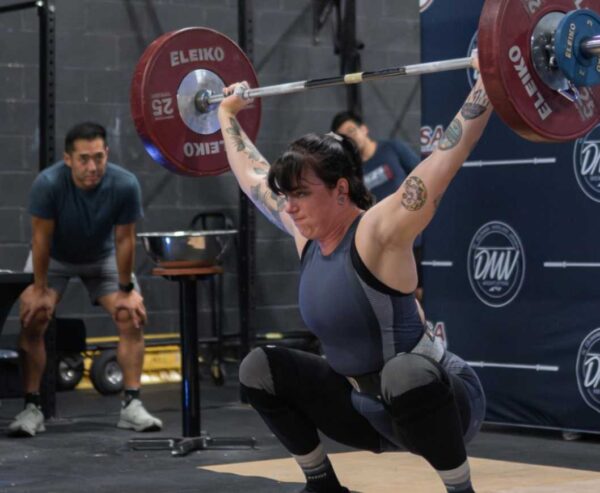 Female weightlifter in a blue singlet overhead squats with a loaded barbell; coach watches in the background at the gym.