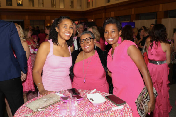 Three women in pink dresses pose together at a pink-themed gala, smiling at a round table with pink décor and drinks nearby.