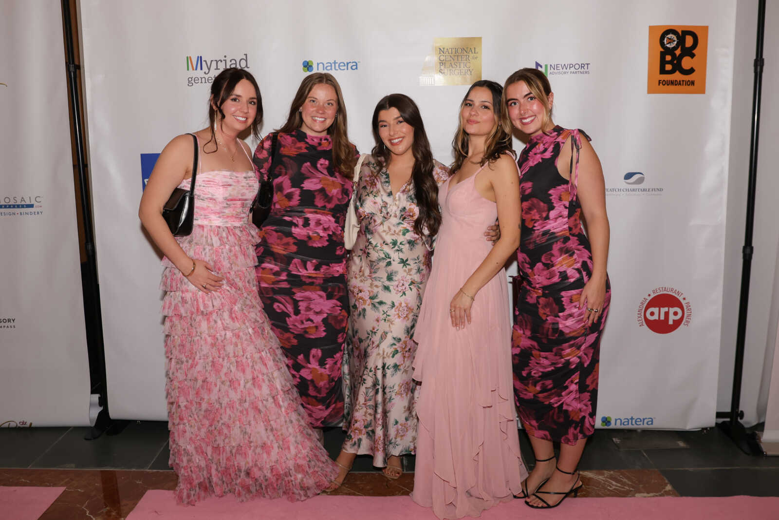 As seen at the ALX Pink Gala (via ALX Pink Gala) | ALXnow Five women in pink and floral dresses pose together on a pink carpet at a formal event, smiling for the photo against a sponsor backdrop.