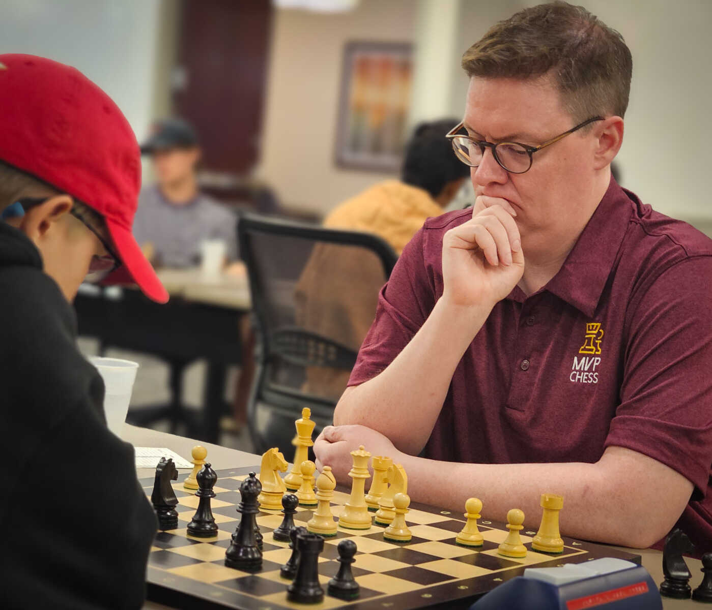 Man with glasses and a maroon polo thinking over a chessboard during a game, pieces arranged in front of him at a table in a cafe-like setting.