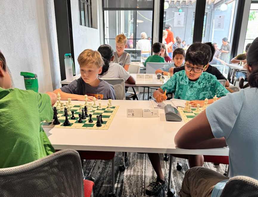 Children seated at a white table playing chess in a bright cafe, with chess clocks and spectators in the background.
