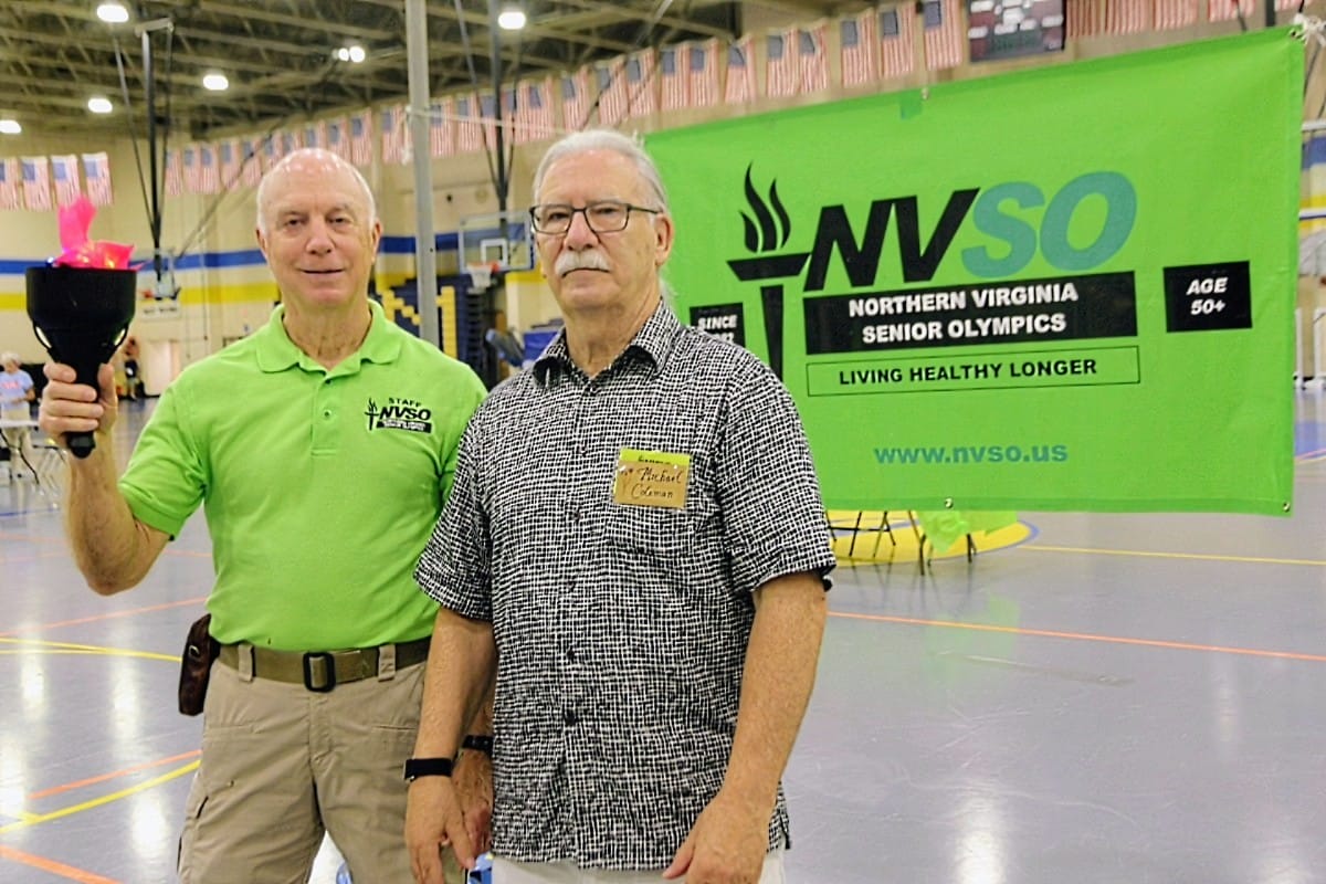 Two older men pose in a gym in front of a bright green NVSO banner; one wearing a lime polo holds a ceremonial torch while the other wears a black‑and‑white patterned shirt with a name badge.