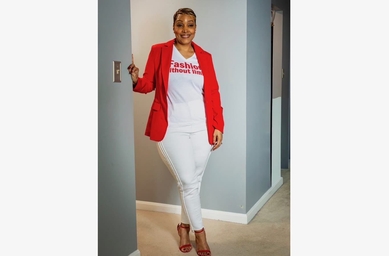 Woman standing in a hallway, wearing a bright red blazer, white outfit, and red sandals; smiling at the camera. The white T-shirt shows the text 'Fashion without limits'.