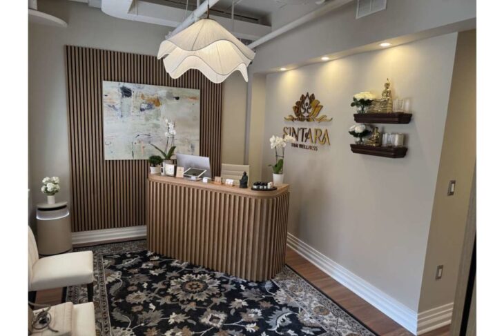 Reception area of a wellness spa with curved wooden front desk, orchid decor, and wall logo reading 'SINTARA Thai Wellness' under warm lighting.