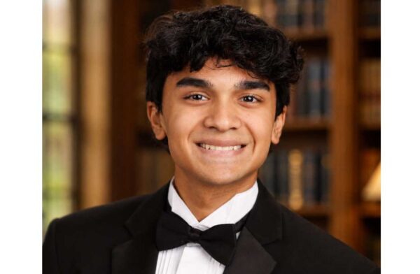 Young man in a black tuxedo and bow tie smiling in a library setting.