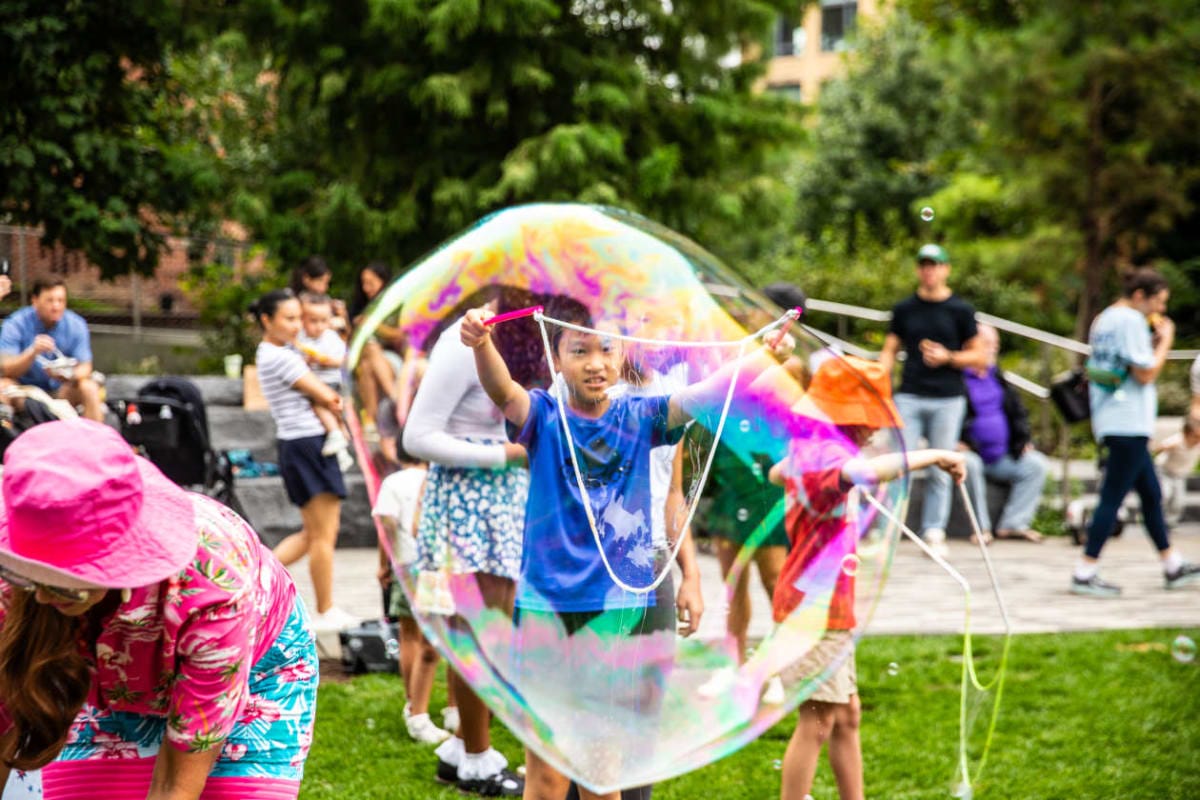 A child in a blue shirt forms a giant rainbow bubble with a wand in a park, with onlookers and green trees in the background.