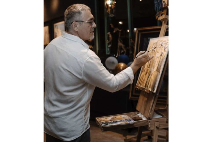 Man in a white shirt painting on a canvas set on an easel in a dimly lit gallery or studio.