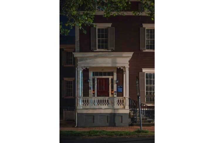 Two-story brick townhouse with a white decorative porch and red front door, lit at night