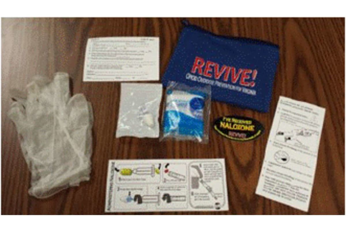 Overdose-reversal kit with blue Revive pouch, naloxone nasal spray, gloves, and printed instructions laid on a wooden surface.