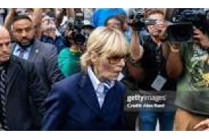 Blond woman in a navy suit walks past a crowd of photographers and reporters taking pictures.
