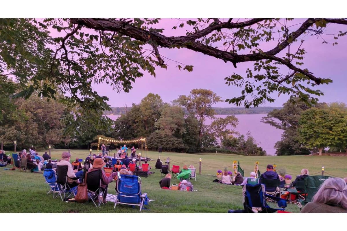 Outdoor lakeside concert with audience seated in folding chairs on the grass under trees at twilight.