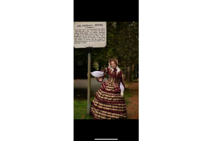 Person in a 19th-century dress posing beside a historical sign that reads 'Lee-Fendall House' in a park setting