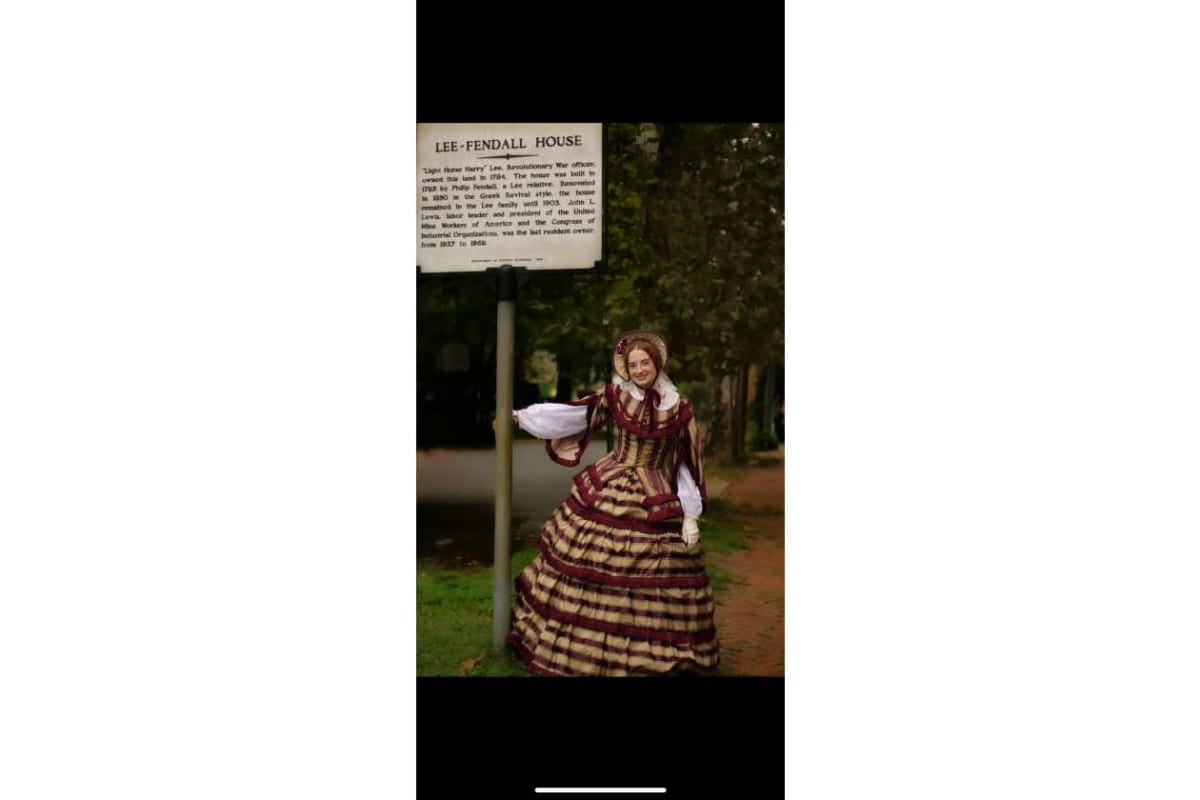 Person in a 19th-century dress posing beside a historical sign that reads 'Lee-Fendall House' in a park setting