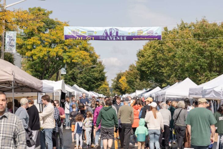 Crowd at an outdoor art fair with white vendor tents lining both sides of a street and a banner overhead reading 'Art on the Avenue'.
