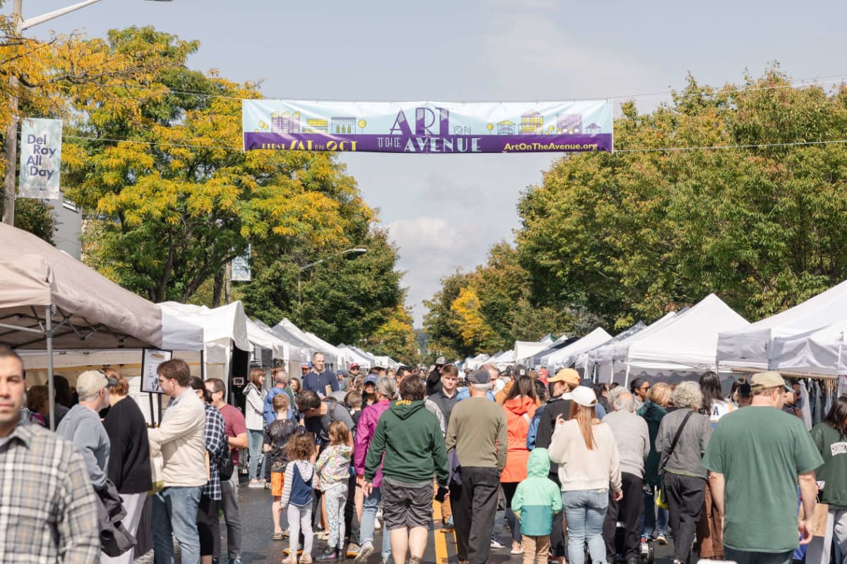Crowd at an outdoor art fair with white vendor tents lining both sides of a street and a banner overhead reading 'Art on the Avenue'.