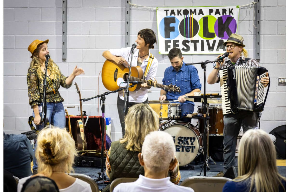 Band performs at Takoma Park Folk Festival: singer with guitar, drummer, and accordionist on stage while audience watches.