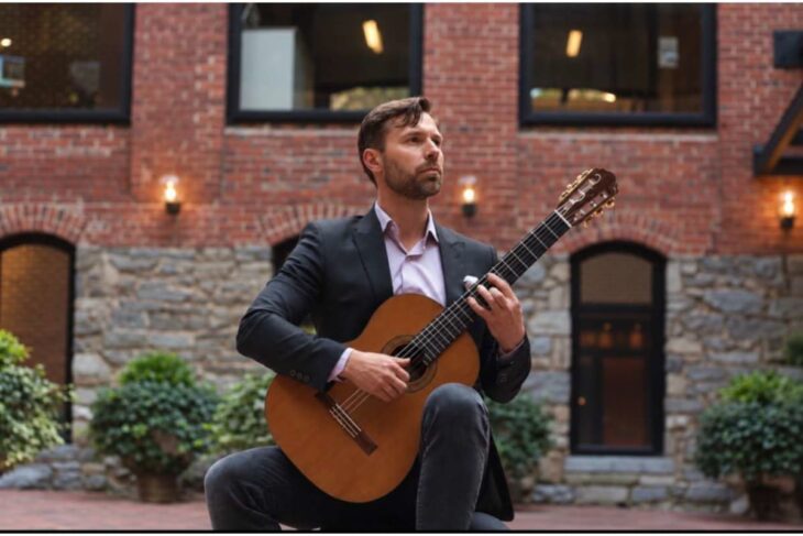 Man in a dark blazer sits and plays a classical guitar in a brick courtyard.