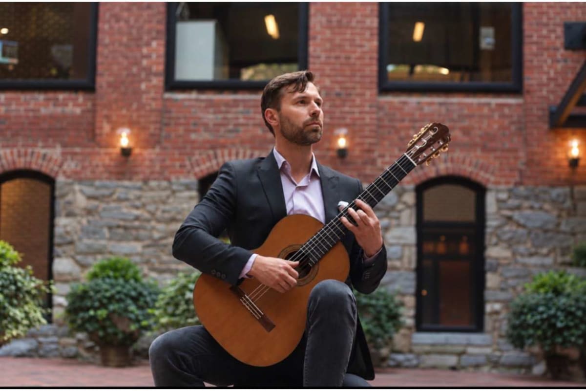 Man in a dark blazer sits and plays a classical guitar in a brick courtyard.