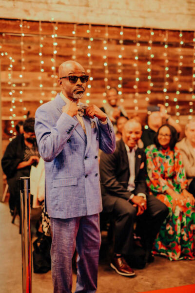 Man in a light blue suit and sunglasses adjusts his bow tie at a formal event, with seated guests and twinkling lights in the wooden backdrop.