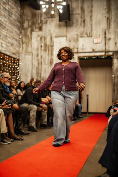 Woman walking a red carpet runway in a maroon cardigan and gray jeans, indoors with audience on both sides.