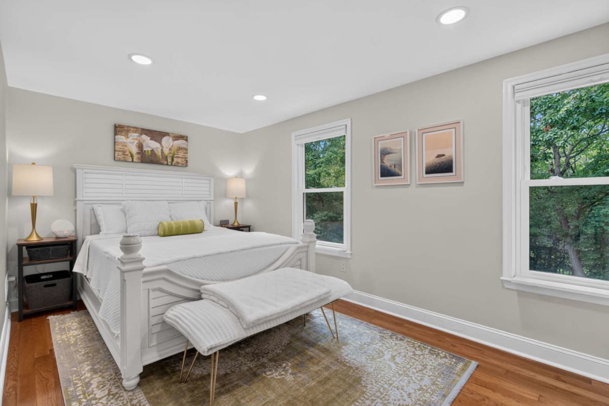 Bright bedroom with a white paneled bed, matching benches, and two nightstands on a warm wood floor; soft beige walls and three window views of greenery on the right.