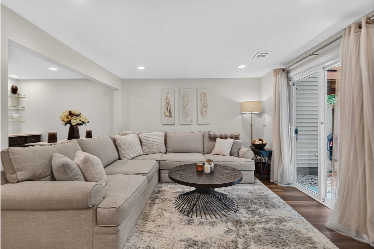 Neutral living room with an L-shaped beige sofa, dark round coffee table, and sliding glass doors with sheer curtains.