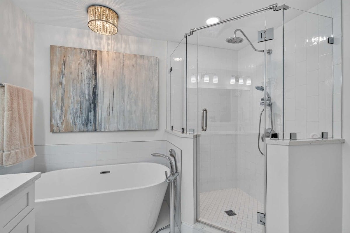 Modern bathroom with a freestanding white tub beside a glass-enclosed shower and an abstract painting on a white-tiled wall.