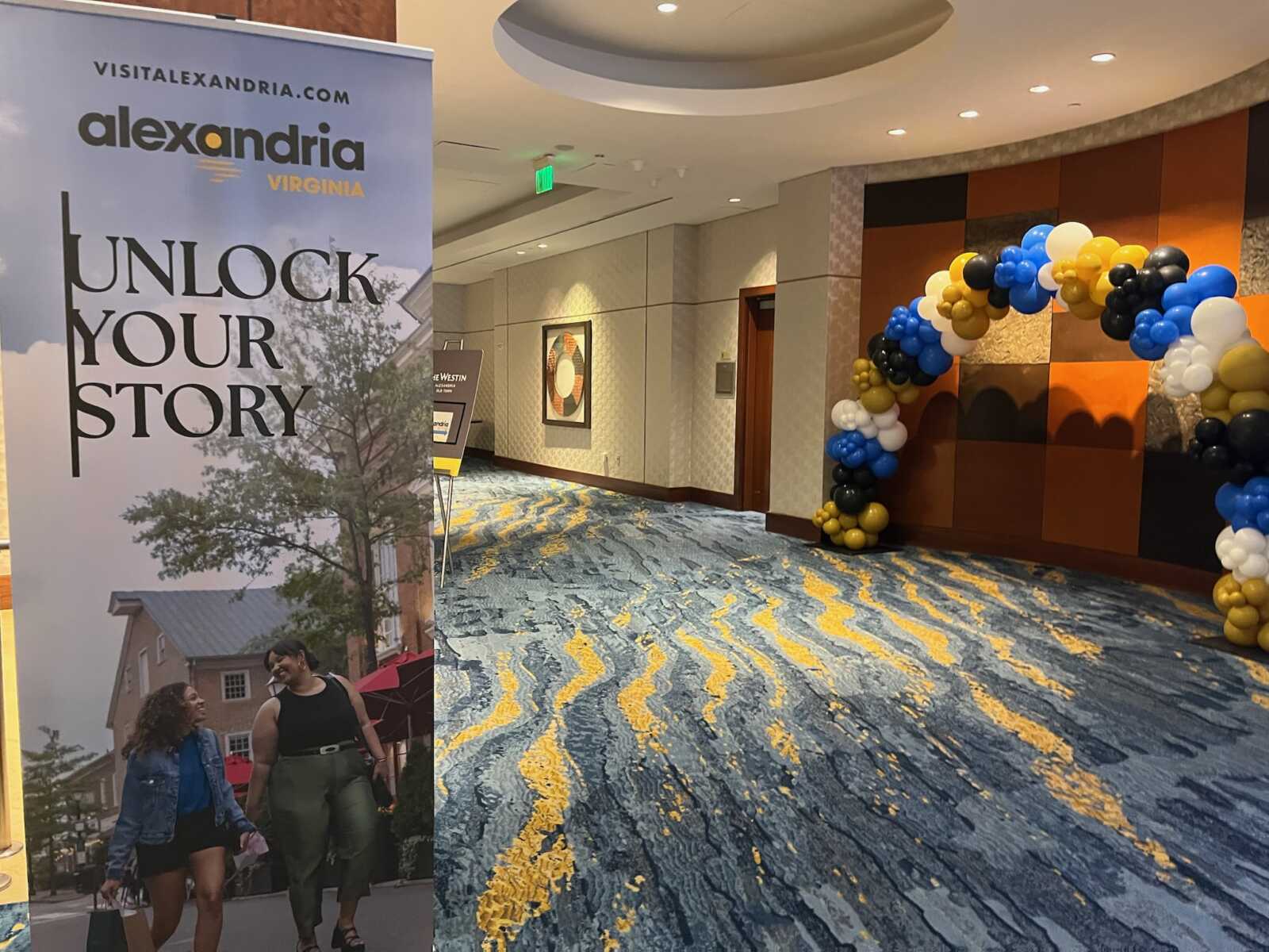 Promotional banner for Alexandria, Virginia with 'UNLOCK YOUR STORY'; two people walking below, and a blue/white/gold balloon arch along a hotel hallway.
