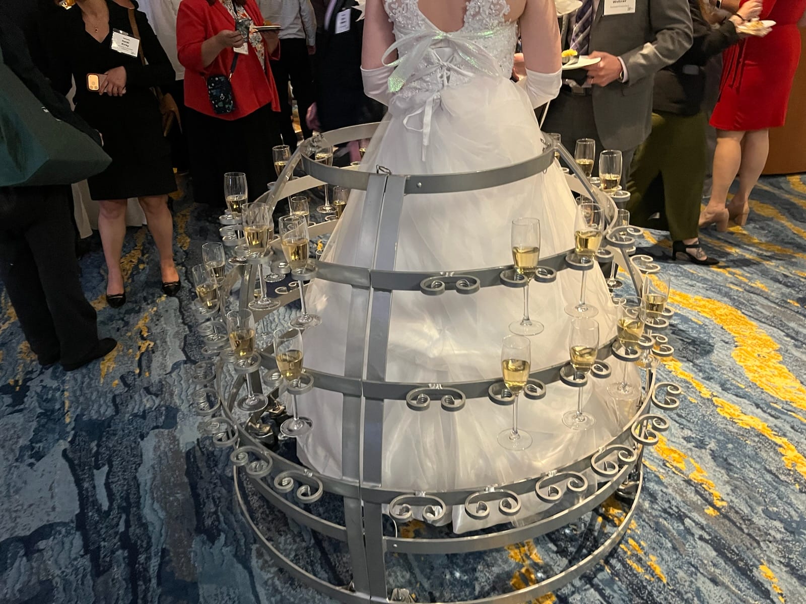 White wedding gown centerpiece surrounded by circular metal racks holding champagne flutes, with guests in formal attire around it.