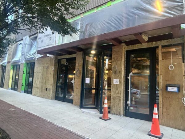 Storefront under renovation with plywood boards on windows, plastic wrap, and orange construction cones on the sidewalk.