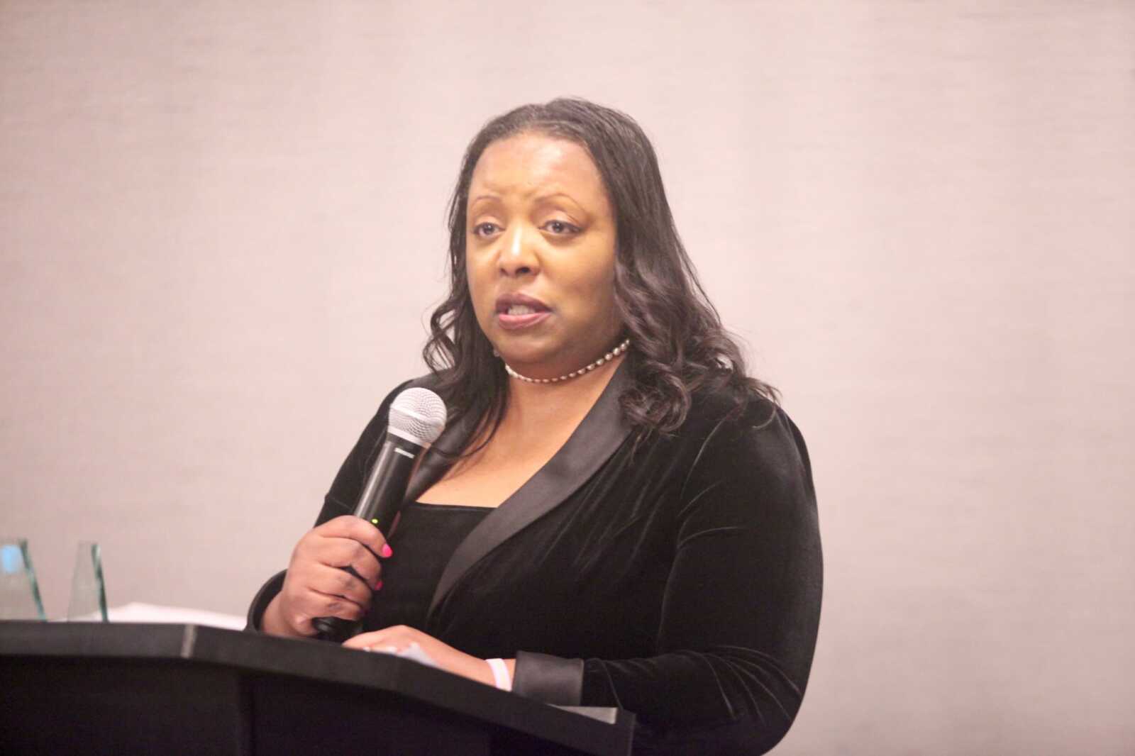 Woman in a black blazer speaking into a handheld microphone at a podium, delivering a speech or presentation.