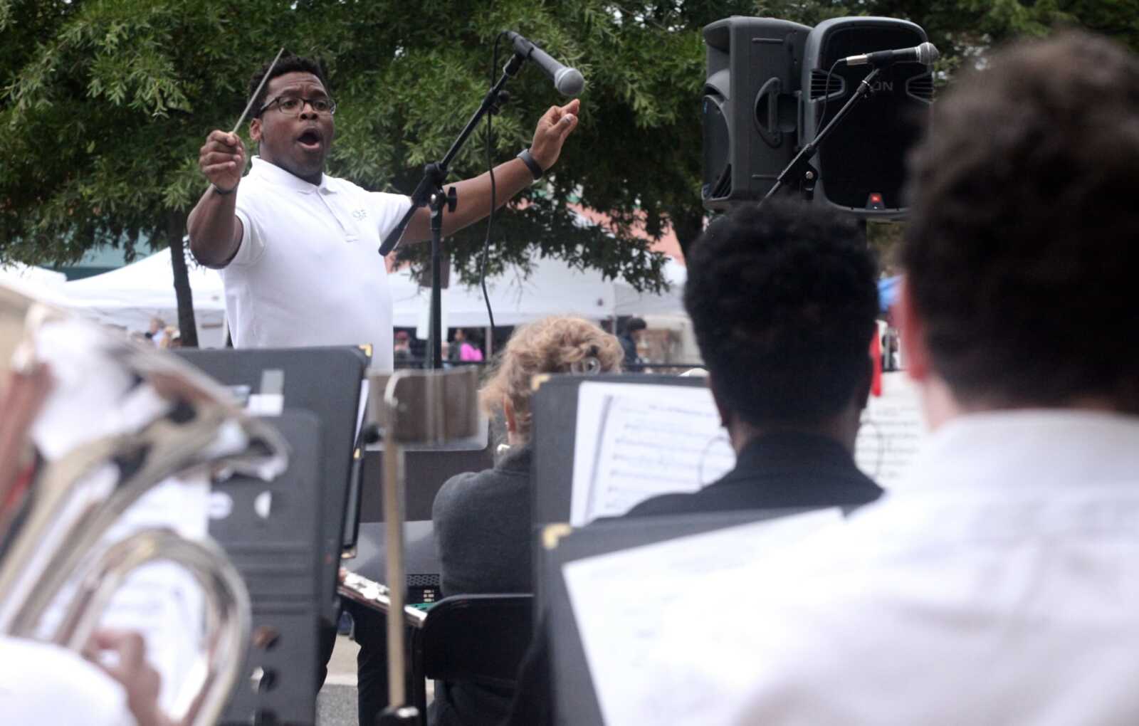 Male conductor in a white polo shirt raises a baton while leading an outdoor band, with a microphone and large speaker nearby and musicians seen from behind.