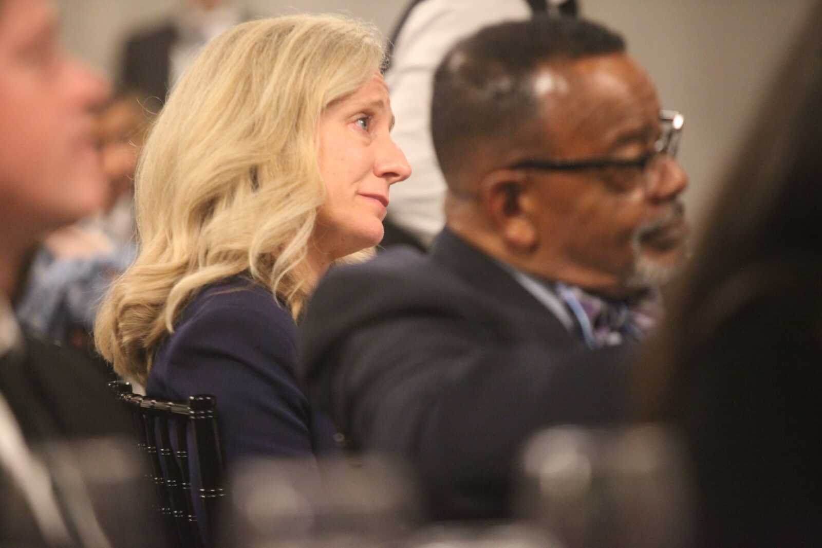 Side profile of a blonde woman wearing a dark blazer, listening at a conference.
