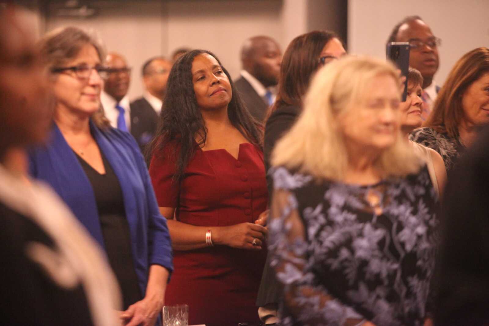 Group of professionally dressed people at a formal event; center woman in a burgundy/red dress looks upward.