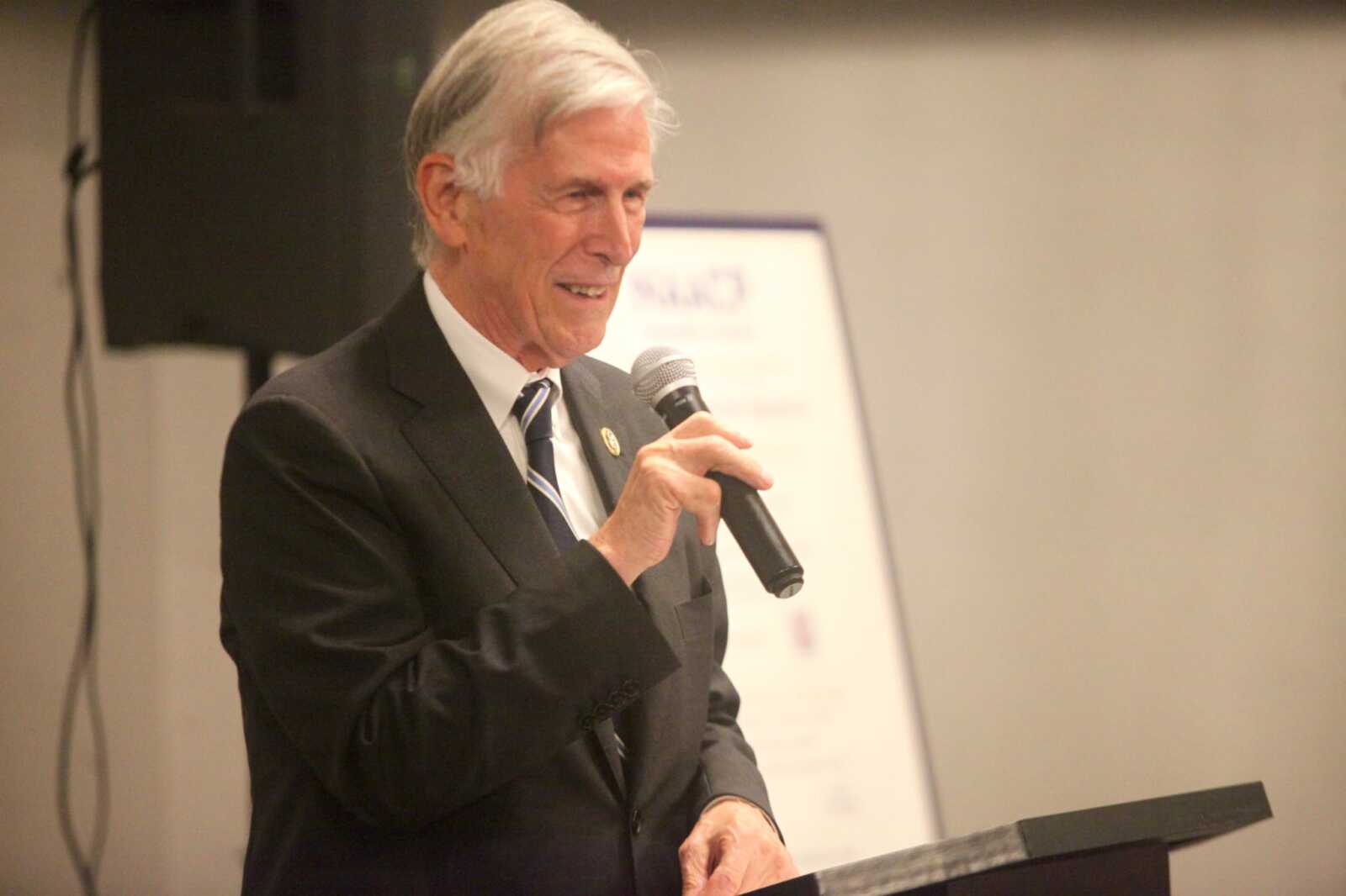 Older man in a dark suit and striped tie speaks into a handheld microphone at a podium during a formal event.