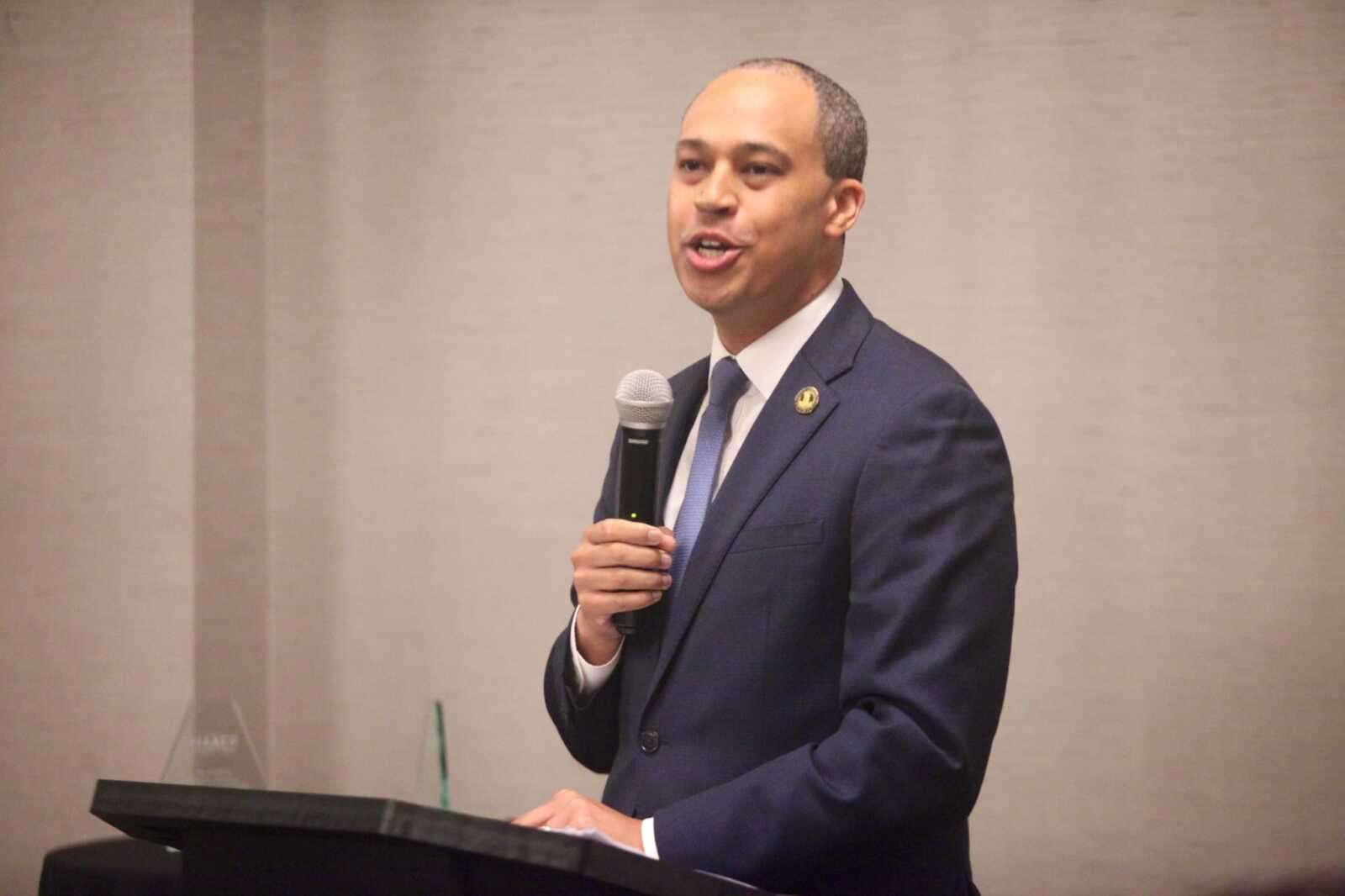 Man in a dark suit and blue tie speaks into a handheld microphone at a podium.