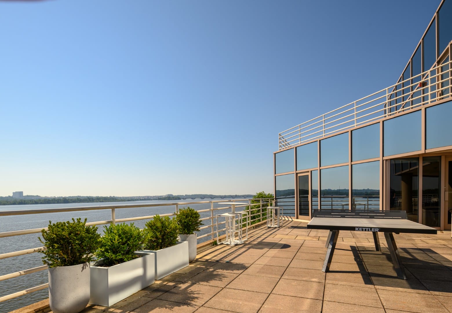 Outdoor terrace along a river with a glass-walled building, railing, and a black Kettler table tennis table on the right, under a clear blue sky.