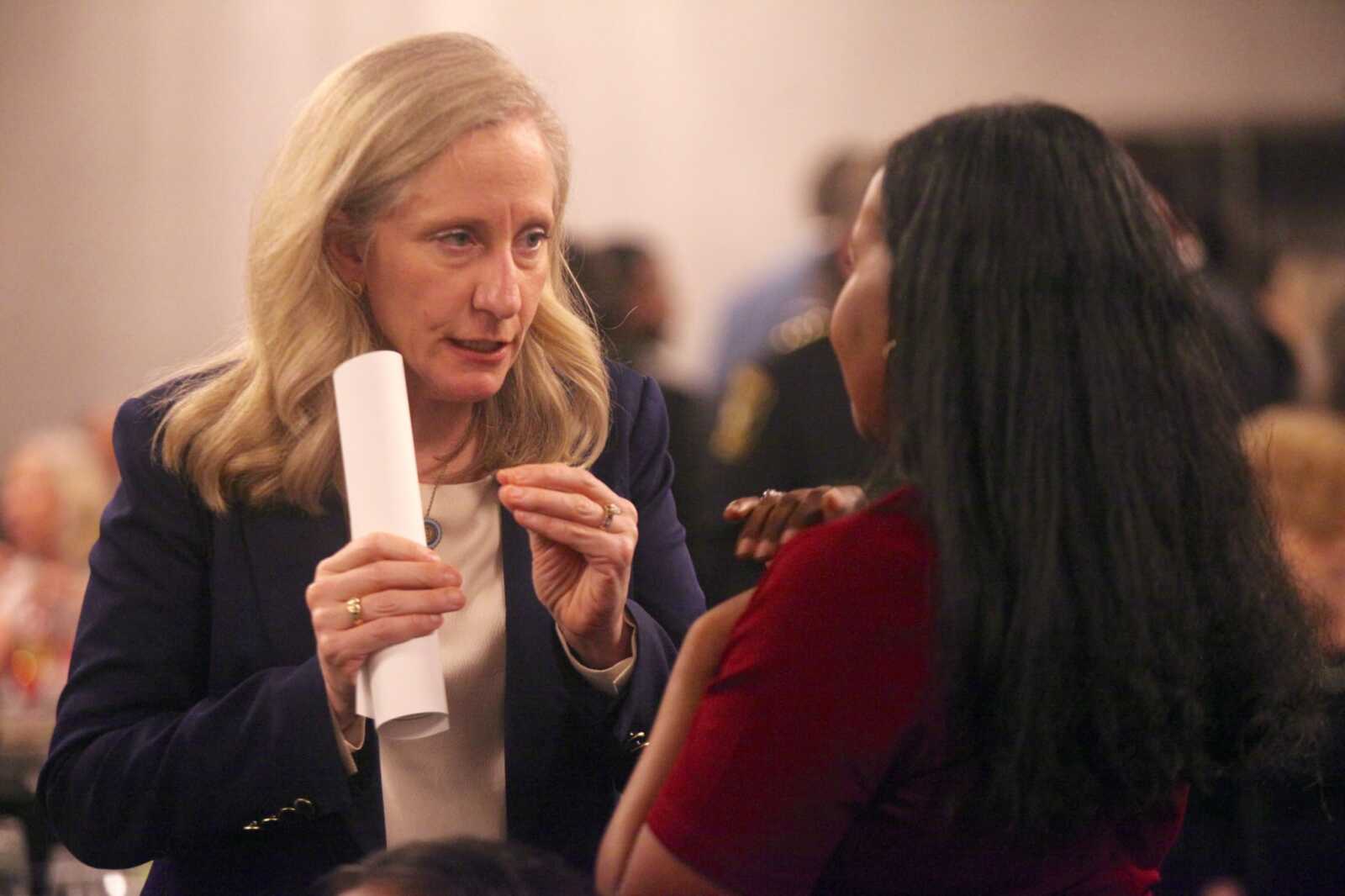 Blonde woman in a navy blazer holds a rolled white document while talking to a woman in a red dress at a formal event.