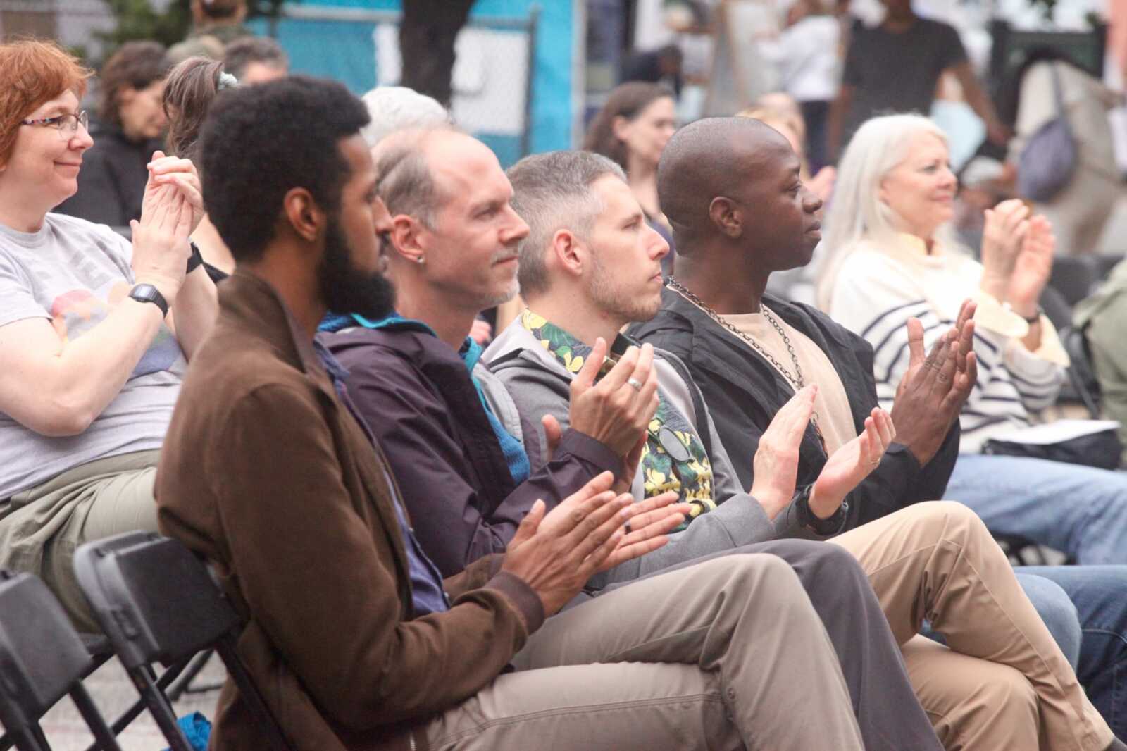 Diverse audience seated outdoors, many people clapping at an event or performance.